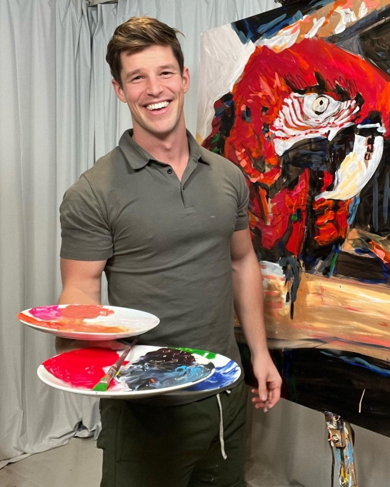 Seb Gower smiling with paint palettes beside a bold parrot painting